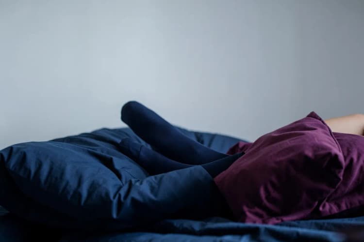 Uma pessoa deitada de costas em uma cama com almofadas extras sob as pernas elevadas, vestindo meias de compressão.