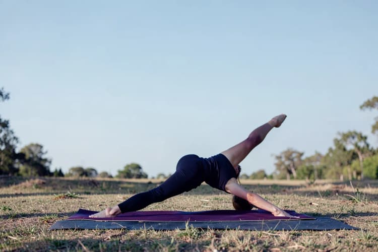 Uma pessoa praticando ioga em uma pose de alongamento de pernas em um ambiente externo com luz natural.