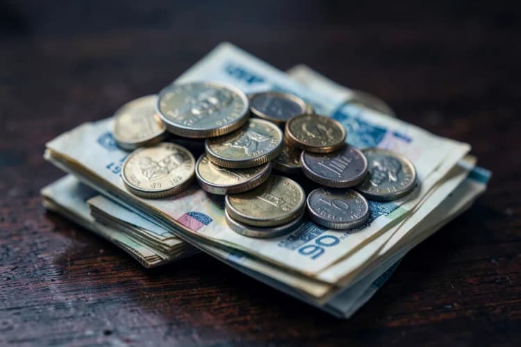 Pilha de moedas de diferentes valores e notas de dinheiro dobradas sobre uma superfície de madeira escura.