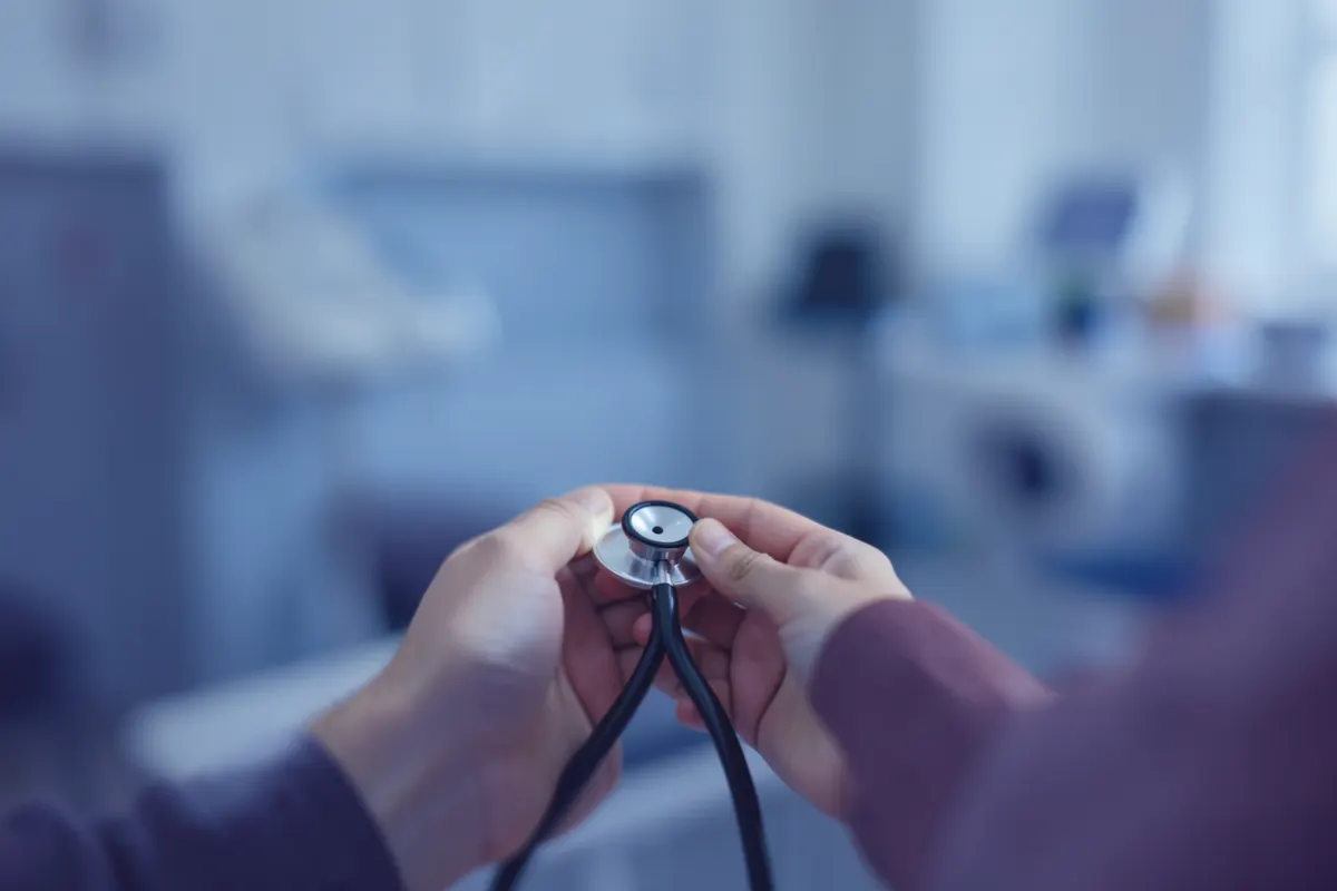 Mãos segurando um estetoscópio sobre um fundo desfocado de consultório médico, em plano médio, com luz natural suave.