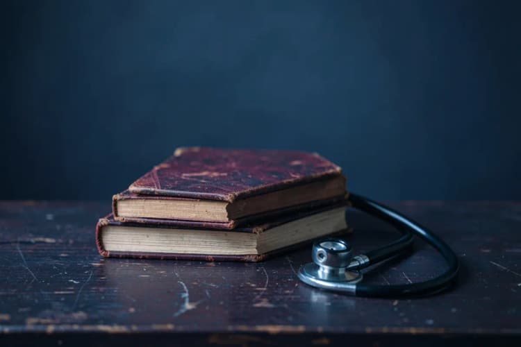 Uma pilha de livros antigos e um estetoscópio sobre uma mesa de madeira escura, sugerindo estudo médico.